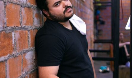 A man in a gym resting against a brick wall, looking tired after exercise.