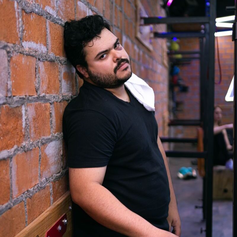 A man in a gym resting against a brick wall, looking tired after exercise.